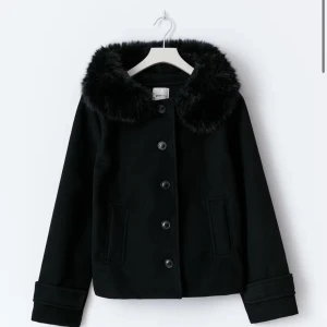 Svart jacka med fuskpäls-krage Gina tricot - Stilren svart jacka med bred fuskpäls-krage och knappar framtill. Jackan har raka ärmar, två fickor och en klassisk, något boxig passform. Perfekt för dig som vill ha en snygg och mysig look under kyliga dagar. Endast använd 1 gång så den är som helt ny ❤️ storlek xs ( köpt för 499)