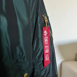 Mörkgrön bomberjacka Alpha Industries - Snygg mörkgrön bomberjacka från Alpha Industries med klassisk 'Remove Before Flight'-tag på ärmen. Jackan har guldfärgade dragkedjor, ribbade muddar och detaljerade fickor. Tillverkad i glansig polyester som ger en clean och modern look.