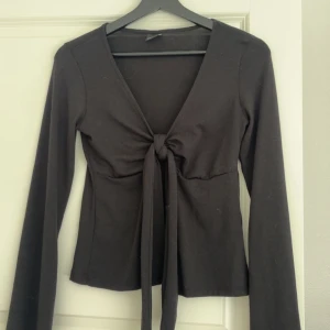 Svart omlottopp från Gina Tricot - Svart långärmad omlottopp från Gina Tricot med knyt framtill och djup v-ringning. Toppens passform är slim och den är tillverkad i mjuk bomullsmix. Perfekt för dig som gillar stilrena och trendiga plagg med snygga detaljer.