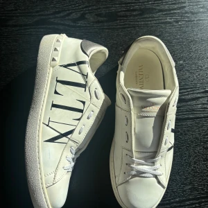Valentino Garavani vita sneakers - Snygga vita sneakers från Valentino Garavani med svart VLTN-logga på sidan och coola nitar runt hälen. Skorna har silvriga detaljer baktill och klassisk snörning. Tillverkade i skinn för en lyxig känsla och stilren look.