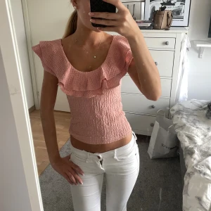 Rosa volangblus med smock - Supersöt ljusrosa blus med smockad kropp och breda volangdetaljer över axlarna. Toppen är croppad och har en rundad urringning. Perfekt för dig som gillar romantisk och feminin stil. Materialet känns lätt och stretchigt.