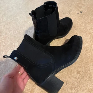 Svarta boots med klack - Snygga svarta boots i mocka och skinnimitation med bred resår på sidorna och dragflik bak. Boots med rund tå och stabil låg klack, perfekta till jeans eller kjol. En liten metallknapp bak ger extra detalj. 🤍