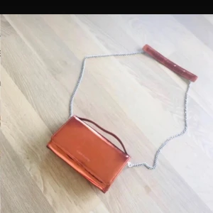 Skimrande väska  - Snygg orange axelväska från Åhléns i glansigt syntetmaterial. Väskan har silverfärgad kedjerem med matchande axelparti och ett stilrent, rektangulärt format. Perfekt för dig som vill sticka ut med en färgstark accessoar. Kolla in mina andra väskor!