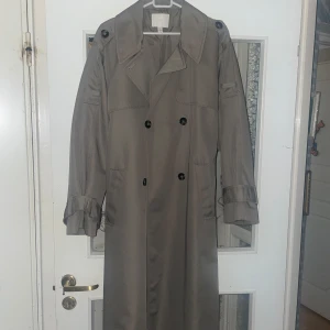 Beige trenchcoat från H&M, storlek M - Säljer en klassisk beige trenchcoat från H&M i storlek M. Jackan har dubbelknäppning, bred krage, knappar på axlarna och bälte i midjan. Långa ärmar med justerbara remmar vid ärmslut. Perfekt för lager-på-lager under vår och höst.