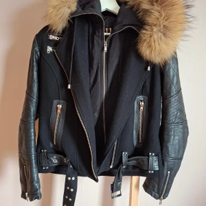 Svart jacka med läderärmar från Bark - Snygg svart jacka från Bark of Sweden med coola skinnärmar och quiltade detaljer. Jackan har en stor pälskrage i brunt, flera dragkedjor, bälte i midjan och långa ärmar. Perfekt höstjacka.  Obs: liten reva på baksidan av ena armen, skulle tro att detta går att fixa för den händige då läder flärpen sitter kvar. Fortfarande mycket kvar att ge och en fin jacka.