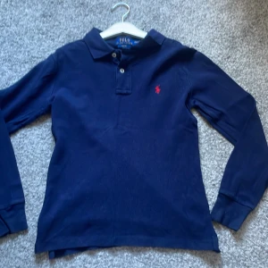 Marinblå långärmad pikétröja Polo Ralph Lauren - Klassisk marinblå långärmad pikétröja från Polo Ralph Lauren i slim fit/10-12. Tröjan har krage, knäppning med två knappar och den ikoniska röda logotypen broderad på bröstet. Perfekt för en stilren look.