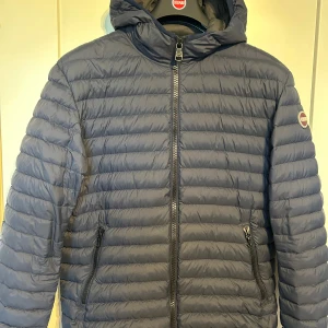 Mörkblå dunjacka från Colmar, strl 52 - Snygg mörkblå dunjacka från Colmar med huva och raka, kvalitet sömmar. Jackan har dragkedja framtill, två fickor med dragkedja och en tydlig Colmar-logga på ärmen. Perfekt för kyliga dagar och riktigt skön att bära. 