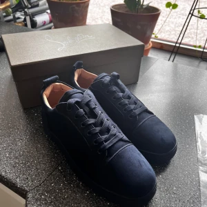 Mörkblå mocka sneakers från Louboutin - Säljer ett par mörkblå sneakers från Christian Louboutin i lyxig mocka med klassisk röd sula. Skorna har rund tå, snörning och beige innersida i skinn. Perfekta för dig som vill sticka ut med exklusiv stil och detaljer.