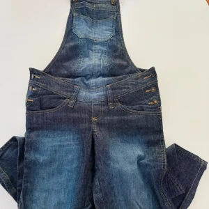 Blå denim hängselbyxor med knappar - Snygga blå denim hängselbyxor (graviditetsbyxor/mamma byxor eg) med klassisk tvätt och slitningar. Byxorna har flera dekorativa knappar i sidorna, framfickor och bakfickor samt justerbara hängslen. Perfekt för dig som gillar en avslappnad och cool stil.