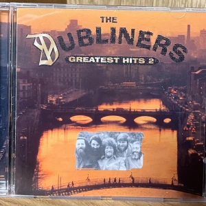 Dubliners - Greatest Hits 2 CD - Upptäck The Dubliners – Greatest Hits 2! En samling klassiska låtar från det legendariska irländska bandet. Perfekt för dig som vill utforska folkmusikens energi och känsla. Fylld med ikoniska låtar och liveinspelningar som passar både nya och gamla fans.