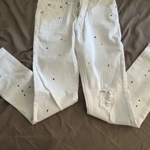 Vita jeans från Purple Brand med stänk - Säljer ett par vita jeans från Purple Brand med svart färgstänk och slitning vid knät. Klassisk femficksmodell i bomull med rak passform. Snygg detalj med silverfärgad patch i midjan. Perfekta för dig som gillar streetwear och unika detaljer.