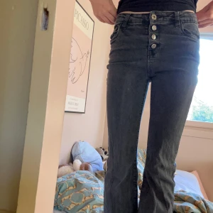 Svarta bootcut jeans med knappar - Snygga svarta jeans med bootcut-ben och hög midja. Byxorna har fyra silvriga knappar framtill, klassiska fickor både fram och bak samt bälteshällor. Materialet är stretchigt jeans och färgen är mörkgråsvart, perfekt för en cool och avslappnad look. Jag är osäker på märket, eftersom att texten på lappen har slitits bort lite. Jeansen är inte nya men knappt använda. Priset kan diskuteras! 