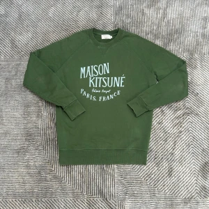 Grön tröja från Maison Kitsuné - Säljer en grön sweatshirt från Maison Kitsuné med vit text på bröstet. Tröjan har rund hals, långa ärmar och ribbade muddar vid ärmslut och nederkant. Perfekt för dig som gillar stilren streetwear med fransk touch.