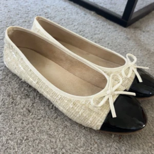 Beige/vita ballerinaskor med svart tå och rosett - Snygga ballerinaskor i beige tyg med glansig svart tå och söt rosettdetalj framtill. Skorna har en klassisk rund tå och platt sula, vilket gör dem både stilrena och lätta att matcha. Perfekta för dig som gillar en chic och tidlös look.