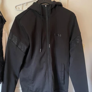 HUGO BOSS hoddie - Svart hoodiejacka från HUGO BOSS med dragkedja och luva. Jackan har diskreta BOSS-loggor på ärmarna och en stilren logotyp på bröstet. Storlek S! Tröjan är äkta går att scanna!