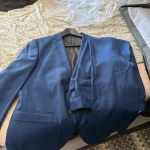 Blå kostym från Esprit - Snygg marinblå kostym från Esprit med klassisk kavaj och matchande kostymbyxor. Kavajen har två knappar, smala slag och flera fickor. Materialet är mjukt och har en lätt glans, perfekt för dig som vill ha en stilren look. Fodret är blått och har Esprit-logga.