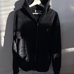 Svart hoodie från A Bathing Ape - Svart hoodie från A Bathing Ape med dragkedja framtill och klassisk huva. Liten BAPE-logga på bröstet och fickor på sidorna. Perfekt för dig som gillar streetwear och vill ha en stilren look. Sitter som L.
