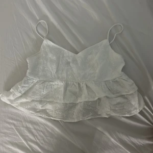 Vit volangig linnetopp med broderi - Supersöt vit linnetopp med tunna axelband och dubbla volanger. Toppen har broderade blommor och är lätt och luftig, perfekt för varma dagar. Snygg v-ringning och romantisk känsla. Passar till jeans eller kjol för en fräsch look.