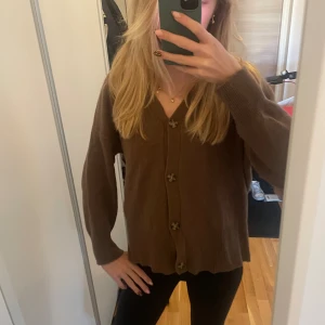 Brun kofta med knappar - Mysig brun kofta med v-ringning och stora dekorativa knappar framtill. Koftan har långärmad passform och är stickad i ett mjukt material, perfekt för lager-på-lager under kyligare dagar.