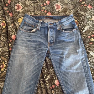 Blå jeans från Nudie Jeans, W30 L34 - Säljer ett par klassiska blå jeans från Nudie Jeans med snygga slitningar och orangea sömmar. Modellen har rak passform och fem fickor. Jeansen är tillverkade i bomull och har en normal midja. Perfekt för dig som gillar tidlös stil.