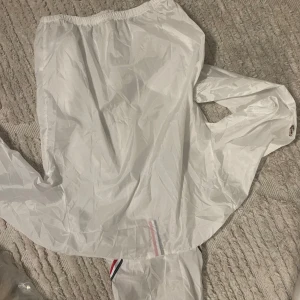 Vit vindjacka från Moncler - Snygg vit vindjacka från Moncler med huva och dragkedja framtill. Jackan har Moncler-logga på ärmen och detaljer i rött, vitt och blått vid fickor och huva. Tillverkad i lätt och glansig polyester, perfekt för blåsiga dagar.