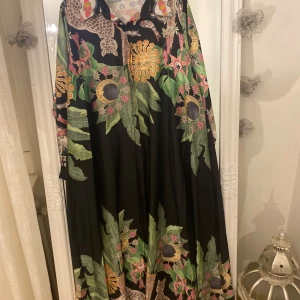 Svart långklänning med tropiskt mönster - Unik svart långklänning med tropiskt inspirerat mönster i grönt, rosa och gult. Klänningen har vida ärmar och är dekorerad med blommor, blad, solar och leoparder. Materialet är tunt och flowy, perfekt för dig som gillar statement-plagg med färg och detaljer.