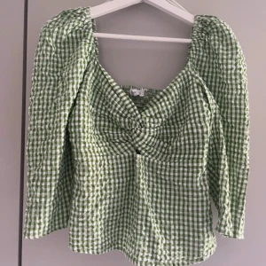 Grön och vit rutig blus med twist - Superfin blus i grönt och vitt med rutigt mönster. Toppen har lång ärm, fyrkantig ringning och snygg twist-detalj framtill. Smockad rygg för extra komfort och volym. Perfekt för dig som gillar en söt och trendig look. Som ny, utan defekter