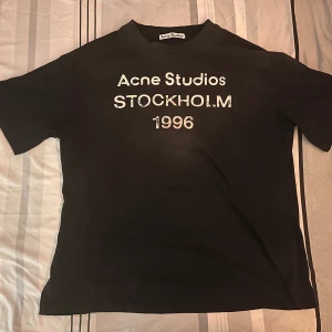 Svart Acne Studios t-shirt Stockholm - Svart t-shirt från Acne Studios med vit text 'Acne Studios STOCKHOLM 1996' på bröstet. Klassisk passform med rund hals och korta ärmar. Tillverkad i mjuk bomull, perfekt för en clean och stilren look. Storlek M 