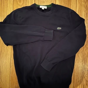 Mörkblå sweatshirt från Lacoste - Snygg mörkblå sweatshirt från Lacoste med klassisk rund hals och det ikoniska krokodilmärket broderat på bröstet. Tröjan har ribbade muddar och en grön detalj på insidan av kragen. Perfekt för en clean och stilren look.