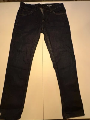 Mörkblå jeans från Tommy Hilfiger - Säljer ett par mörkblå jeans från Tommy Hilfiger med klassisk femficksdesign och gul kontrastsöm. Jeansen har rak passform och diskret logga på bakfickan. Materialet är slitstarkt jeans och modellen är tidlös och enkel att styla.