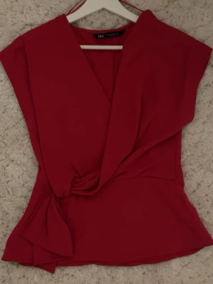 Röd omlottopp från Zara XS - Säljer en snygg röd omlottopp från Zara i storlek XS. Toppen har en v-ringad design och knyt i midjan som ger en cool och feminin look. Materialet är mjukt och lätt, perfekt för att styla med jeans eller kjol. Färgen är intensivt röd och modellen är ärmlös.