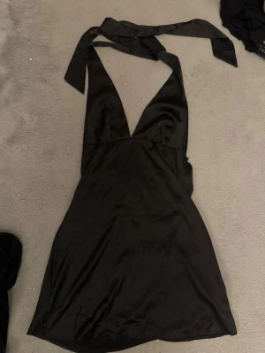 Svart omlott kortklänning med knyt - Snygg svart kortklänning i satin med omlott-design och djup v-ringning. Klänningen har breda band som knyts runt halsen och en slät, glansig yta. Perfekt för dig som gillar stilrena och trendiga plagg med lite edge.