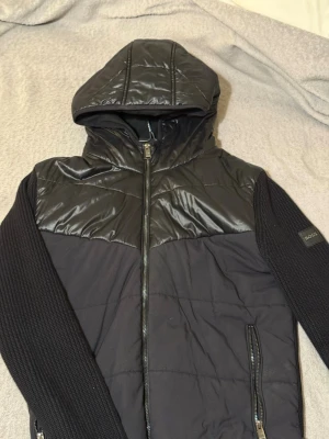 Svart pufferjacka med stickade ärmar från BOSS - Svart pufferjacka från BOSS med glansig huva. . Ärmarna är stickade och har en BOSS-logga på vänster arm. Jackan har dragkedja framtill och två sidofickor. Ny pris runt 3000