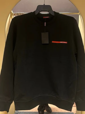 Svart sweatshirt från Prada - Svart sweatshirt från Prada med rund hals och lång ärm. Diskret röd logga på bröstet ger en clean och stilren look. Tröjan har ribbade muddar vid ärmslut och nederkant. Perfekt för dig som gillar exklusiva märken och enkel design.
