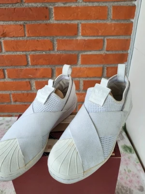 Adidas Superstar Slip-On vita sneakers 38 - Säljer ett par Adidas Superstar Slip-On sneakers i helvitt med meshpaneler och breda elastiska band över foten. Skorna har klassisk rund tå med gummiskal, Adidas-logga på plösen och hälflik för enkel påtagning. Snygg och modern design som sticker ut.