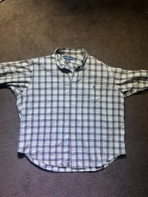 Rutig skjorta från Ralph Lauren XL - Säljer en klassisk rutig skjorta från Ralph Lauren i storlek XL. Skjortan är i 100% bomull och har en snygg färgkombo med grått, vitt, beige och röda linjer. Den har långa ärmar, knappar framtill och den ikoniska broderade loggan på bröstet.
