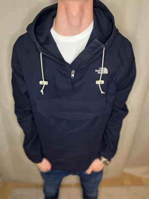 Mörkblå vindjacka från The North Face - Snygg mörkblå vindjacka från The North Face med huva och vita snören. Jackan har en half zip framtill och en stor ficka på magen. Perfekt för dig som gillar streetwear och vill ha en stilren look. Skick 8/10. Vi kan gå ner i pris vid snabb affär. // Studio II Resell