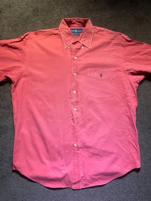 Röd skjorta från Ralph Lauren M - Säljer en snygg röd skjorta från Ralph Lauren i storlek M. Skjortan är tillverkad i 100% bomull och har klassisk krage, knappar framtill och en bröstficka med blå broderad logga. Perfekt för dig som gillar stilrena och tidlösa plagg.