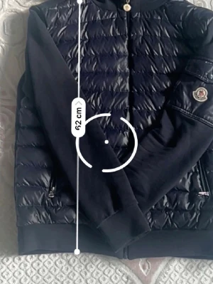 Svart pufferjacka från Moncler - Svart pufferjacka från Moncler med huva och quiltad framsida. Jackan har dragkedja, långärmade släta ärmar och Moncler-logga på armen. Materialet är en mix av polyester och syntet, vilket gör den både lätt och varm. Perfekt för dig som vill ha en snygg och sportig look.