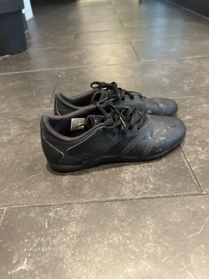Adidas Predator svarta fotbollsskor - Säljer ett par svarta Adidas Predator fotbollsskor med snyggt diskret mönster och meshpaneler på sidorna. Skorna har snörning och platt sula, perfekt för inomhusspel. Materialet är syntet och mesh, och designen är stilren med svarta detaljer.