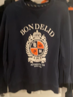 Mörkblå sweatshirt från Bondelid - Snygg mörkblå sweatshirt från Bondelid med stort tryck och emblem i vitt och orange på bröstet. Tröjan har rund hals och långa ärmar, tillverkad i mjuk bomull. Perfekt för dig som gillar klassisk och sportig stil.