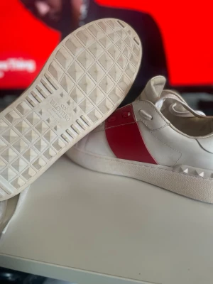 Valentino Garavani vita sneakers med röd detalj - Säljer ett par snygga Valentino Garavani sneakers i vitt skinn med bred röd panel på sidan och coola nitar runt hälen. Skorna har rund tå, platt sula och klassisk snörning. Perfekt för dig som vill ha en lyxig och unik look. Har inte skolådan eller kvitto