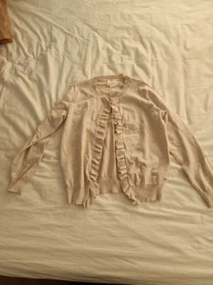 Beige kofta med volang från Holly & Whyte - Snygg beige kofta från Holly & Whyte by Lindex med volangdetaljer längs öppningen framtill. Koftan har rund hals, långa ärmar och ribbade muddar. Perfekt att slänga på sig när det är lite kyligt ute.