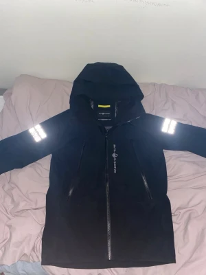 Svart Sail Racing jacka med huva - Svart jacka från Sail Racing med huva och reflekterande detaljer på ärmarna. Jackan har två långa dragkedjor framtill och meshfoder på insidan. Materialet är 100% polyester och jackan är designad för att tåla tuffa väder. Perfekt för dig som gillar att vara ute oavsett väder.