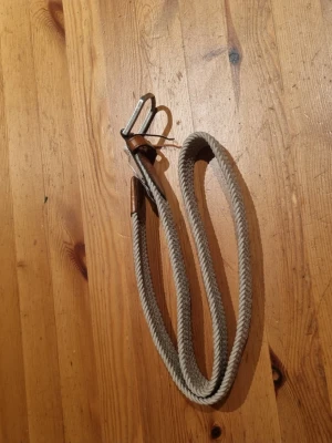 Beige flätad bälte med läderdetaljer - Säljer ett snyggt beige flätat bälte med bruna läderdetaljer vid spännet och änden. Bältet har ett klassiskt metallspänne och är gjort i ett slitstarkt syntetmaterial som ger en cool och avslappnad vibe. Perfekt för att lyfta din outfit.