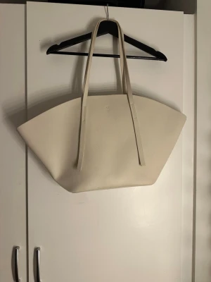 Beige shoppingväska i skinn - Stor och stilren shoppingväska i beige skinn med minimalistisk design och långa handtag. Väskan har en unik, bred form som gör den perfekt för att bära med sig mycket grejer. Passar dig som gillar enkel och modern stil.