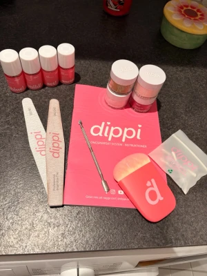 Nagelkit med färgade dip powders - Nagelkit från Dippi med fyra dip powders i färgerna grön, röd, ljusrosa och nude samt tillhörande vätskor. Inkluderar två nagelfilar, en metallisk nagelverktyg och en mjuk borste med rosa handtag. Perfekt för att skapa snygga och kreativa naglar hemma.