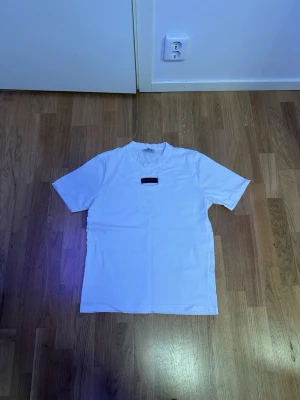 Vit Burberry t-shirt med logga - Säljer en clean vit t-shirt från Burberry i storlek M. Klassisk rund hals och korta ärmar. På bröstet sitter en svart patch med Burberry-loggan i silver. Tillverkad i mjuk bomull för skön känsla hela dagen.
