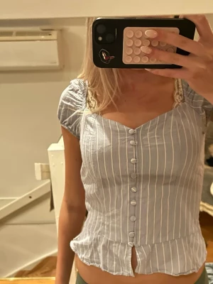 Blåvit randig blus med spetsdetaljer - Säljer en söt blus i ljusblått med vita vertikala ränder och dekorativa knappar framtill. Blusen har korta puffärmar, spetsdetaljer vid axelbanden och en liten slits nedtill. Materialet känns lätt och somrigt, perfekt för varmare dagar. Skulle säga att det är storlek xs/ s