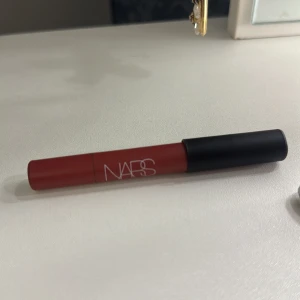 NARS  läppstift-penna - Säljer en snygg läppstift-penna från NARS i en matt rosa röd nyans. Pennan har en smidig form som gör det enkelt att applicera färgen exakt på läpparna. Hylsan är röd med svart lock och vit NARS-logga. Perfekt för dig som gillar intensiva färger och vill ha ett trendigt resultat.